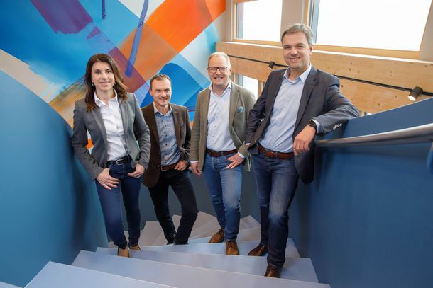 [Translate to Deutsch:] Our 4 members of management are pictured on a modern blue staircase, with a large colorful mural in the background.